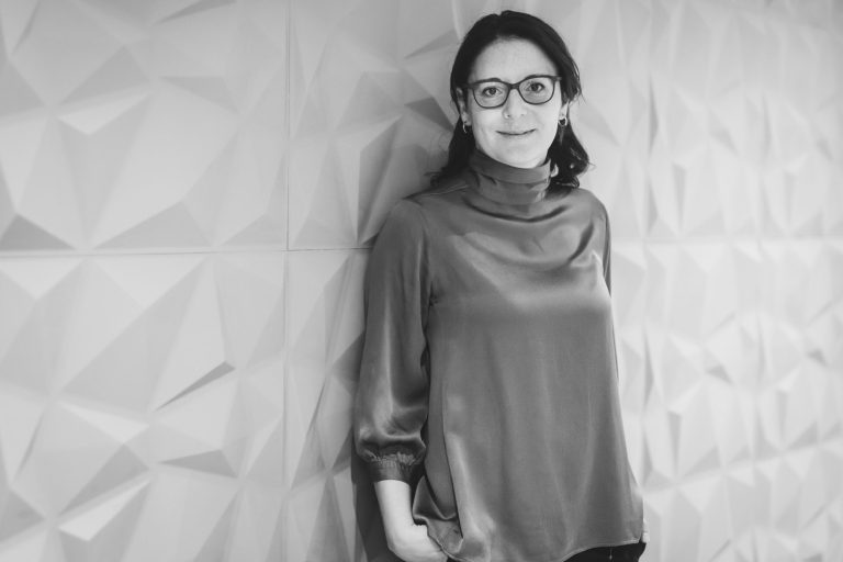 Frau mit Brille in einem modernen, geometrischen Raum, lächelt entspannt.