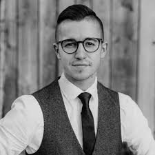 Mann in einem Anzug mit Krawatte, Brille und kurzem Haarschnitt vor einer Holzwand.