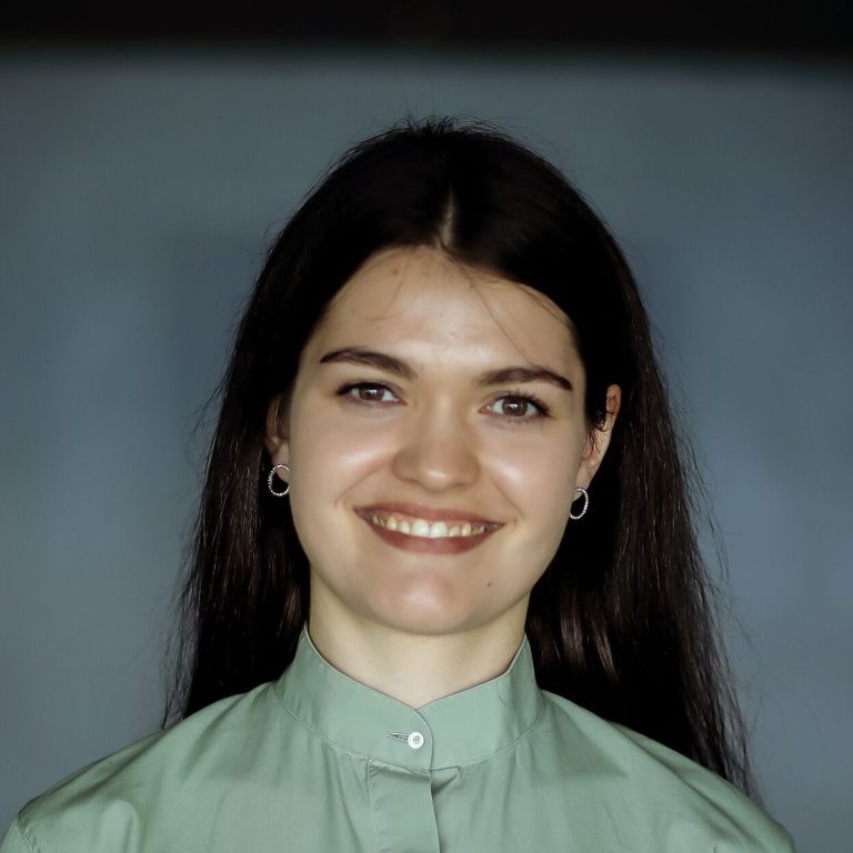 Junge Frau mit langen, dunklen Haaren und freundlichem Lächeln, trägt ein grünes Oberteil.